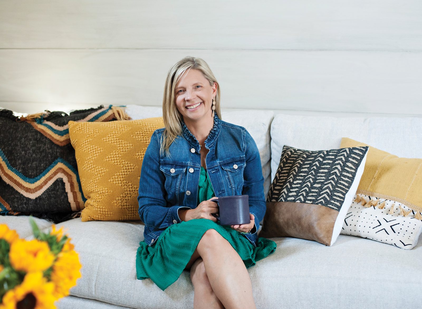 Julie Gates relaxing on a couch with a coffee mug and sunflowers nearby — a warm, welcoming portrait.
